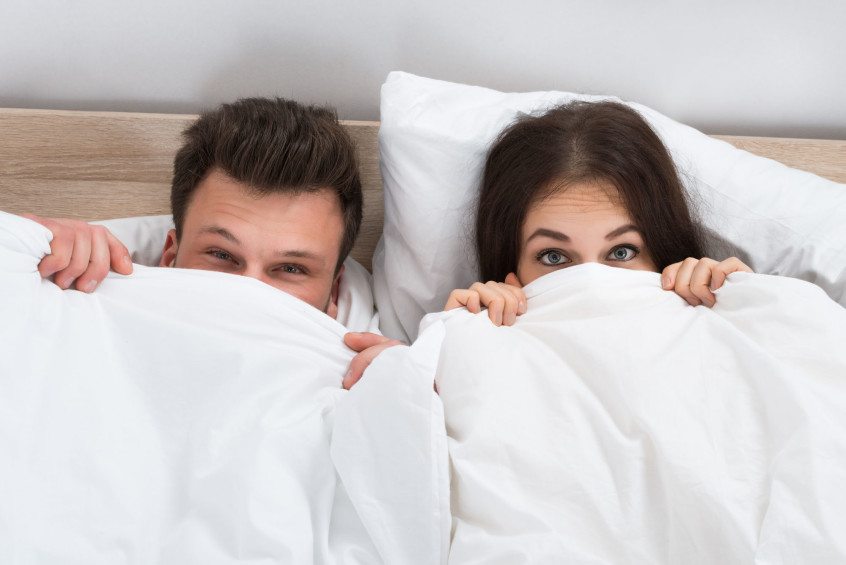 A man and woman hiding under the covers in bed.