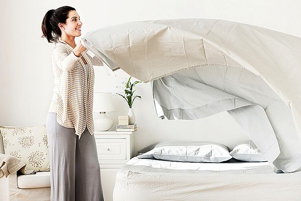 A woman is holding up sheets in front of her bed.