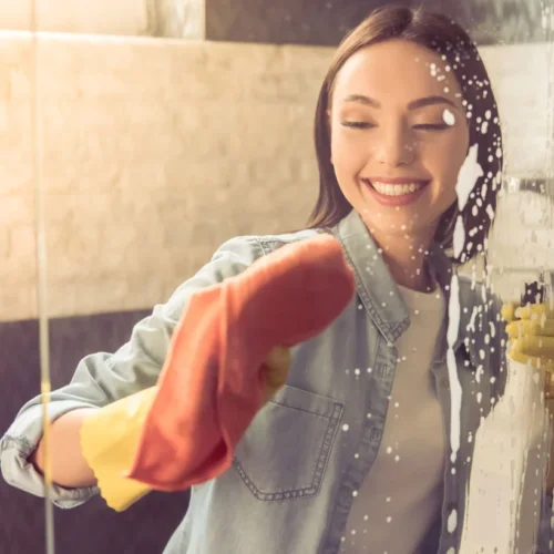 How-to-Clean-Soap-Scum-Off-Glass-Shower-Doors- A woman holding a spray bottle and cleaning the shower.
